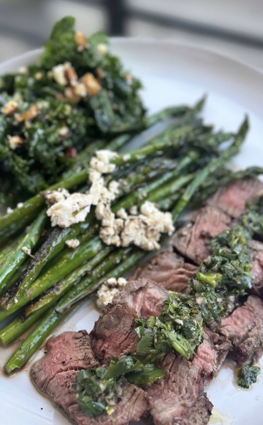 Sliced steak with herb sauce, asparagus, and greens on a white plate